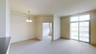 This single-floor space presents a clean, understated design centered around an open layout with a neutral color palette throughout. The main living area is finished with plush wall-to-wall carpeting in a soft gray tone, contributing to a calm and comfortable atmosphere. Cream-colored walls and white ceilings enhance the sense of brightness, while crown molding adds a subtle architectural detail that frames each room with a classic touch.

A pendant chandelier with a traditional multi-arm design serves as a focal point, providing both ambient lighting and a decorative accent in the main living area. Large vertical-blind-covered windows along one wall allow considerable natural light to filter into the space, contributing to its open and airy feel.

An interior doorway connects the main living area to an adjoining room, which also features carpeting and its own window, maintaining consistent design continuity throughout the space. The layout appears straightforward and functional, with clearly defined zones that offer versatility for various uses.

Overall, the space presents a well-maintained, neutrally designed interior that provides a bright and livable environment.