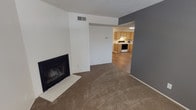 This single-floor space is thoughtfully laid out, with a living area and kitchen that flow naturally into one another. The living room is finished with wall-to-wall carpeting in a neutral tan tone, providing a warm and comfortable foundation. A fireplace with a tile hearth and dark surround serves as a focal point on one wall, adding both a functional and visual anchor to the room. The walls are painted in a clean white, with one accent wall in a deep charcoal gray, creating a modern contrast. A textured popcorn ceiling runs throughout, and a ceiling-mounted HVAC vent ensures year-round climate control.

An open passageway connects the living area to the kitchen, which features hard surface flooring in a wood-tone finish. The kitchen is equipped with light oak cabinetry offering ample storage, white appliances including a range and microwave, and a practical, efficient layout suited for everyday cooking. Under-cabinet space and countertops provide functional workspace. The overall ambiance of the space is clean and move-in ready, with a straightforward, functional design throughout.