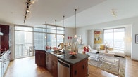 This well-appointed 2-bedroom, 2-bathroom high-rise residence offers an open, single-floor layout filled with natural light from expansive floor-to-ceiling windows and glass walls that frame sweeping city skyline views. The open-concept kitchen is a standout feature, showcasing rich dark wood cabinetry, granite countertops, a stone tile backsplash, stainless steel appliances including double wall ovens and a French door refrigerator, and a curved center island with bar seating illuminated by pendant lighting. The kitchen flows seamlessly into the dining area and living room, where large windows provide panoramic urban views and access to a private balcony. The primary bedroom features tray ceilings, a ceiling fan, plush carpeting, and a large window with notable city views. Its en-suite bathroom includes a soaking tub, glass-enclosed walk-in shower with stone tile surround, dual vanity with granite countertops, and tile flooring. The second bedroom is equally well-lit with city views and soft carpeting. A dedicated laundry room with side-by-side washer and dryer adds everyday convenience. Hardwood floors run throughout the main living areas.