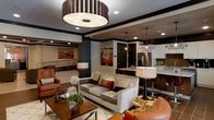 This single-floor space showcases a thoughtfully designed open-concept layout that seamlessly connects the living area to the kitchen, creating a fluid and inviting environment. The living room is anchored by a plush gray sofa and complemented by rich cognac leather accent chairs, with decorative throw pillows in warm terracotta tones adding a pop of color. A striking large drum pendant light with a slatted wood surround serves as a dramatic focal point overhead, while recessed lighting throughout ensures a well-lit atmosphere. Dark-toned wall trim and ceiling details frame the space with a bold, contemporary aesthetic. A wooden coffee table styled with organic décor elements, including dried botanicals and greenery, grounds the seating area with a natural touch. Bold abstract artwork adorns the walls, adding depth and visual interest. The adjacent kitchen features dark cabinetry, stainless steel appliances, a stone countertop island with bar seating, and elegant pendant lighting above. The overall ambiance is sophisticated and modern, blending warm earth tones with darker architectural elements to create a space that feels both refined and welcoming.