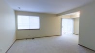 This two-floor space offers a clean, functional layout designed to maximize comfort and usability. The main living area, pictured here, is a generously proportioned room finished with wall-to-wall plush carpeting in a neutral tone, contributing to a warm and quiet atmosphere. The room is illuminated by natural light entering through a large horizontal window fitted with white blinds on one wall, while a sliding glass door with vertical blinds on the adjacent wall provides both additional light and direct access to an outdoor area. The neutral beige wall tones throughout create a versatile backdrop suitable for a variety of interior styles. Dark wood baseboard trim runs along the base of the walls, adding a subtle but grounding contrast to the light color scheme. The open floor plan of this level allows for flexible furniture arrangements, making the space adaptable to different living needs. The two-floor configuration of the overall property suggests additional living, sleeping, or utility space on the upper or lower level, expanding the functional capacity of the home well beyond what is visible in this primary living area.