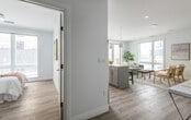 This thoughtfully designed single-floor residence showcases a clean, contemporary aesthetic throughout, unified by light hardwood flooring, crisp white walls, and an abundance of natural light streaming through large windows fitted with horizontal blinds. The open-concept layout connects the living, dining, and kitchen areas seamlessly, creating an inviting flow ideal for everyday living and entertaining. The kitchen is a standout feature, offering gray shaker-style cabinetry, white quartz countertops, a subway tile backsplash, stainless steel appliances, and a generous island with bar seating beneath modern pendant lighting. The living area is bright and airy, with oversized windows and access to a balcony that frames views of the surrounding urban neighborhood. Two well-proportioned bedrooms each benefit from large windows that invite ample daylight, with one featuring a floor-to-ceiling window overlooking the streetscape. The bathroom is finished with marble-patterned tile flooring, a glass-enclosed walk-in shower with white subway tile surround, and a clean vanity. Throughout the residence, recessed lighting complements the natural light, and the consistently neutral color palette lends the space a polished, modern character.