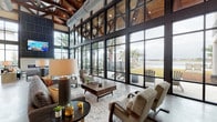 This sophisticated single-story clubhouse seamlessly blends modern industrial and rustic design elements to create a dynamic, welcoming communal space. The centerpiece of the building is its dramatic great room, where soaring ceilings with exposed wood beam trusses and visible industrial ductwork rise above polished concrete floors. A sweeping wall of steel-framed, floor-to-ceiling windows and glass doors floods the interior with natural light while offering unobstructed views of the outdoor grounds, palm trees, and a waterfront beyond.

The grand main entrance features a double-height glass façade with black steel framing, creating an impressive and transparent arrival experience. Throughout the space, carefully curated lounge seating areas invite gathering and relaxation, with plush sofas, accent chairs, and coffee tables arranged atop layered area rugs. An accent wall clad in reclaimed wood adds warmth and texture, complementing the industrial aesthetic.

Separate, more intimate seating nooks offer a quieter retreat, furnished with upholstered accent chairs, patterned rugs, and decorative wall art, with large windows providing views of the surrounding neighborhood. A glass-walled conference or leasing room provides a sleek, transparent workspace visible from the main floor. The overall ambiance is refined yet approachable, balancing raw architectural elements with warm, thoughtfully designed interiors.