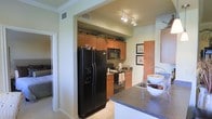This single-floor residence presents a well-appointed kitchen as its central feature. The kitchen is outfitted with a full suite of black appliances, including a large side-by-side refrigerator with an in-door ice and water dispenser, a built-in microwave, and a gas range with an overhead exhaust. Warm-toned wood cabinetry extends to the upper walls, providing generous storage, and is topped with decorative greenery that adds a touch of natural detail. The dark granite countertops offer ample prep space and carry a polished, durable aesthetic throughout. Track lighting mounted on the ceiling delivers targeted illumination across the workspace. A kitchen island or peninsula with matching granite extends into the foreground, enhancing both functionality and the sense of openness. Light-toned tile flooring runs through the kitchen area, contributing to a clean and bright atmosphere. Framed artwork on the adjacent wall adds a decorative accent, while a hallway visible beyond the kitchen hints at additional living spaces. The overall ambiance is practical yet refined, with cohesive finishes creating a unified and welcoming environment.