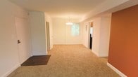 This single-floor space offers a clean and functional layout with a bright, open feel throughout. The main living and dining area is finished with warm beige carpeting that provides a cozy underfoot experience, while crisp white walls and white baseboards give the interior a fresh, well-maintained appearance. A decorative chandelier serves as a focal point in the dining area, adding a touch of elegance to the otherwise understated design. A large window allows natural light to flow generously into the space, offering a pleasant view of the surrounding greenery outside. The transition from carpeted areas to hardwood flooring near the entryway adds a subtle contrast in materials, defining different functional zones within the open layout. Multiple doorways lead to additional rooms, suggesting a well-connected and versatile floor plan. The overall ambiance is light, airy, and neutral, making it an inviting and adaptable space suited to a variety of uses and personal styles. The single-floor design ensures easy accessibility and a seamless flow between rooms throughout the home.