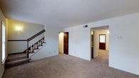 This two-storey space in Summit offers a straightforward and functional layout across both levels. The main floor features a generously sized living area finished with neutral wall-to-wall carpeting, creating a clean and understated aesthetic throughout. A staircase with a dark wood banister and white balustrade runs along one wall, providing access to the upper floor while adding a classic architectural detail to the space. The main living area opens toward a hallway that connects to additional rooms, with a closet door visible along the corridor. Further down the hall, a wood-paneled entry door with a window allows natural light to filter into the space, brightening the passage. A glimpse of what appears to be a kitchen or utility area is visible adjacent to the hallway. Overhead HVAC venting is integrated into the ceiling, contributing to the practical nature of the layout. The white walls and light ceiling throughout the main floor maintain a neutral, versatile canvas. Overall, the space presents a clean, move-in ready environment with a traditional residential feel, well-suited to a variety of uses across its two floors.