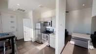 This single-floor unit offers a thoughtfully designed open-concept layout that maximizes functionality and flow. The kitchen is a standout feature, equipped with stainless steel appliances including a refrigerator, electric range, and over-the-range microwave, complemented by gray shaker-style cabinetry, stone-look countertops, and a classic subway tile backsplash. Dark hardwood-style flooring runs throughout the main living areas, grounding the space with warmth and continuity. A partial dividing wall separates the kitchen from the living area, which provides ample room for seating and additional furnishings. Natural light pours into the bedroom through multiple windows, including tall vertical panes that brighten the crisp white walls. The bedroom offers a serene, minimalist ambiance with enough space for a full-sized bed and storage. The bathroom is clean and well-appointed, featuring a tub-shower combination with white tile surround, a marble-look countertop vanity, and brushed nickel fixtures. Additional doors suggest a closet or storage area near the entry. Overall, the unit presents a modern, neutral aesthetic with practical design choices suited for comfortable everyday living.