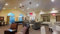This single-floor community clubhouse offers a welcoming and well-appointed space designed for both relaxation and entertainment. The main lounge area is anchored by soaring vaulted ceilings that create an open, airy atmosphere, complemented by a combination of recessed lighting and stylish pendant fixtures, including a bold red drum shade that adds a pop of color. Comfortable seating arrangements featuring plush gray upholstered chairs and accent tables are thoughtfully arranged throughout, encouraging conversation and leisure. Large arched windows allow natural light to flow into the billiards area, which is equipped with a full-size pool table, adding a recreational focal point to the space. Adjacent to the main lounge, a bar or kitchenette area with counter seating provides a convenient space for refreshments and gatherings. Neutral tones in warm beige and gray run throughout the walls and flooring, creating a cohesive and inviting aesthetic. An elevator is also visible, ensuring accessible access across the facility. Overall, the space balances sophistication with functionality, making it an ideal setting for community events, casual socializing, or quiet relaxation.