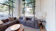 This single-floor professional space strikes an ideal balance between comfort and functionality, offering a refined environment suited for both collaborative work and focused meetings. The layout is anchored by a stylish lounge area furnished with a modular sectional sofa in a neutral gray fabric, complemented by layered textiles, a woven area rug, and a round wood coffee table. Sheer white curtains frame the oversized, black-framed floor-to-ceiling windows that run along the perimeter, flooding the interior with abundant natural light and providing open views of mature street trees and the surrounding neighborhood streetscape. Warm-toned hardwood flooring runs throughout, lending continuity and a sense of warmth to the space. Adjacent to the lounge, a small conference room features built-in connectivity at a clean white meeting table surrounded by contemporary rounded barrel chairs. The room is distinguished by rich teal-blue accent walls, curated wall art, a whiteboard for collaborative use, and wall-mounted sconce lighting that adds a polished finish. A second, more compact meeting room mirrors the functional aesthetic with its own white conference table, dark seating, and direct window views to the outdoors. Together, the areas create a cohesive, light-filled workspace that feels both professional and inviting.