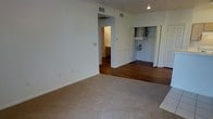 This single-story space is designed with a practical and straightforward layout. The main living area features neutral-toned carpeting that provides a comfortable underfoot feel, while the adjoining spaces transition to rich, dark hardwood flooring, creating a clear visual distinction between zones. Crown molding runs along the ceiling, adding a subtle architectural detail, while recessed lighting and a ceiling vent ensure adequate illumination and climate control throughout.

An open doorway leads to a bathroom, which is partially visible and features wood cabinetry, suggesting functional storage. Adjacent to this, a utility area is accessible through a set of bifold doors, housing what appears to be a water heater and laundry hookups, offering added convenience. The walls throughout are painted in a clean white tone, contributing to a bright and neutral aesthetic. A thermostat is mounted on the wall, indicating individual climate control. The overall layout is efficient, with each area flowing naturally into the next, making the space functional for everyday living.