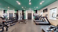 Located at 36 Stony Hill Road in Bethel, CT, this fitness center is a thoughtfully designed exercise space that delivers a comprehensive workout environment within a compact, efficient footprint. The room is immediately distinguished by its bold teal ceiling, which contrasts sharply with the white walls adorned with motivational graffiti-style lettering — including phrases like "Mindset Is Everything," "Believe," and "Achieve" — creating an energizing atmosphere from the moment you enter.

The flooring consists of durable rubber-speckled gym material, ideal for high-impact activities and equipment placement. A full-length mirror spans one wall, visually expanding the space and allowing users to monitor their form during workouts. Ceiling fans and recessed lighting ensure the room remains well-ventilated and brightly lit throughout.

The cardio section is equipped with multiple treadmills, an elliptical trainer, and a stationary bike, positioned near windows that allow natural light to filter in. The strength-training area features a multi-station cable machine, a full dumbbell rack with adjustable benches, and additional weight equipment. Mounted flat-screen TVs provide entertainment options during workouts. The space is clean, modern, and purposefully laid out to maximize usability across both cardio and strength disciplines.