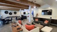 This single-floor communal space blends rustic charm with contemporary design, creating an inviting and versatile environment for socializing and relaxation. The ceiling is a standout architectural feature, showcasing bold exposed wooden beams that add warmth and character to the open layout. Soft white walls provide a clean backdrop that allows the curated décor to take center stage.

The main lounge area is anchored by plush, neutral-toned sofas accented with vibrant coral throw pillows, arranged around a sturdy reclaimed wood coffee table. Pendant globe light fixtures hang overhead, casting a warm, ambient glow throughout the space. A built-in counter area with a mounted flat-screen TV offers a cozy nook, while a sculptural horse head figurine and a large colorful art piece add artistic focal points.

Further into the space, a billiards table is surrounded by intimate seating arrangements with dark-toned chairs and tables, ideal for casual gatherings. An additional wall-mounted television enhances the entertainment options in this zone. The light wood-toned flooring ties the entire space together, complementing both the rustic beam ceiling and the modern furnishings. Overall, the space exudes a sophisticated yet welcoming ambiance suited for community and leisure.