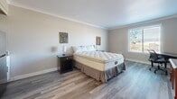 Located at 2886 S Circle Dr in Colorado Springs, CO, this efficiently designed studio unit offers a well-organized single-floor layout that maximizes every square foot. The main living area serves as both bedroom and living space, featuring light gray wood-look laminate flooring, neutral-toned walls, and elegant crown molding that adds a refined architectural touch throughout. Natural light filters in through a window fitted with blinds, brightening the open room. The space accommodates a king-sized bed, two nightstands, a dedicated work desk with an ergonomic chair, and a media console — providing a comfortable and functional environment. A compact kitchenette is tucked into one corner of the main room, outfitted with white cabinetry, a microwave, a sink, and a tiled backsplash, offering convenient in-unit food preparation. The bathroom features light ceramic tile flooring, a vanity with a granite-style countertop, and a full bathtub and shower combination complete with a safety grab bar and a curtained enclosure. The entrance flows naturally into the main living area, contributing to the unit's cohesive and practical design. Overall, the space presents a clean, neutral aesthetic suited for comfortable single-occupancy living in Colorado Springs.