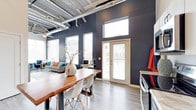 This single-floor space makes a strong design statement through its industrial-chic aesthetic, combining raw architectural elements with contemporary finishes. The open-plan layout seamlessly connects the dining and living areas, creating a fluid and functional living environment.

The ceilings soar overhead, showcasing exposed silver ductwork and mechanical systems that contribute to the space's urban industrial character. Deep charcoal-black walls provide a dramatic backdrop throughout, contrasting boldly with light hardwood-style flooring that runs continuously across the space.

Natural light floods the interior through multiple windows fitted with white horizontal blinds, including transom windows positioned high on the walls to maximize brightness. A set of French doors with a transom above opens to an outdoor area, further enhancing the sense of openness and connectivity to the exterior.

The dining area features a rich, solid wood live-edge table paired with modern white Eames-style chairs, striking a balance between warmth and contemporary design. Adjacent to this, the living area offers a comfortable seating arrangement with colorful accent cushions. The overall ambiance is bold, modern, and thoughtfully curated.