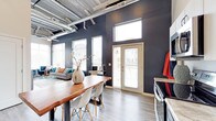 This single-floor space makes a strong design statement through its industrial-chic aesthetic, combining raw architectural elements with contemporary finishes. The open-plan layout seamlessly connects the dining and living areas, creating a fluid and functional living environment.

The ceilings soar overhead, showcasing exposed silver ductwork and mechanical systems that contribute to the space's urban industrial character. Deep charcoal-black walls provide a dramatic backdrop throughout, contrasting boldly with light hardwood-style flooring that runs continuously across the space.

Natural light floods the interior through multiple windows fitted with white horizontal blinds, including transom windows positioned high on the walls to maximize brightness. A set of French doors with a transom above opens to an outdoor area, further enhancing the sense of openness and connectivity to the exterior.

The dining area features a rich, solid wood live-edge table paired with modern white Eames-style chairs, striking a balance between warmth and contemporary design. Adjacent to this, the living area offers a comfortable seating arrangement with colorful accent cushions. The overall ambiance is bold, modern, and thoughtfully curated.