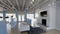 This contemporary single-floor space showcases an open, airy design with exposed white-painted steel trusses and recessed track lighting that add an industrial-chic character to the interior. The soaring ceiling height creates a sense of expansiveness throughout the main area. Large windows and glass French doors along one wall invite abundant natural light, offering views of the outdoor surroundings and seamlessly connecting the interior to the outside.

The lounge area is anchored by a clean-lined white sofa and a modern square coffee table, set atop a dark textured area rug over light hardwood-style flooring. A wall-mounted flat-screen television is prominently positioned above a floor-level stone fireplace, which serves as a striking focal point with its natural stone surround and raised hearth.

Adjacent to the lounge, a sleek kitchenette features white cabinetry, a countertop with a sink, and bar-height seating, making it ideal for casual gatherings and events. The walls are finished in a cool blue-grey tone, complementing the white structural elements and light flooring throughout. Together, these features create a welcoming, versatile space well-suited for socializing and community use.