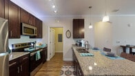This single-story residence is thoughtfully designed with a cohesive aesthetic throughout, combining dark espresso cabinetry, granite countertops, and wood-style flooring to create a warm, modern atmosphere. Crown molding accents the main living and kitchen areas, adding a refined architectural detail.

The kitchen is a functional centerpiece, featuring rich dark cabinetry extending to the ceiling, stainless steel appliances including a refrigerator, range, and over-the-range microwave, and granite countertops. A large peninsula with pendant lighting provides additional prep and seating space, while track lighting ensures excellent task illumination.

The living room is generously proportioned, with dark hardwood-style flooring, crown molding, a ceiling fan, and large windows that allow natural light to fill the space. The open layout connects seamlessly to surrounding areas.

The bedrooms feature plush carpeting, ample natural light from well-positioned windows, and access to adjacent bathrooms. Each bathroom is finished consistently with dark vanity cabinetry, granite countertops, wood-style flooring, and a tiled tub-shower combination with marble-look tile surrounds, offering a clean and cohesive design.