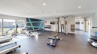 Located on Idlewild Road in Bel Air, Maryland, this single-level fitness center offers a comprehensive workout environment designed to accommodate a wide range of exercise needs. The space is thoughtfully organized into distinct functional zones, allowing users to move seamlessly between cardio training, strength work, and free-weight exercises.

The cardio area is well-appointed with multiple treadmills and elliptical machines, each equipped with individual monitors for an enhanced workout experience. Large windows along the perimeter flood the space with abundant natural light while providing views of the surrounding outdoor greenery, creating an inviting and energizing atmosphere. Wall-mounted televisions are positioned throughout for added convenience.

The strength training section features a cable machine system, a dedicated dumbbell rack with a full range of weights, and multiple adjustable benches to support varied workout routines. A standout design element is the bold geometric accent wall rendered in dark charcoal and teal, adding a modern, dynamic aesthetic to the space.

The fitness center is finished with durable rubber flooring throughout, recessed lighting in the white ceiling, and a built-in water fountain station. The open floor plan and generous spacing between equipment ensure a comfortable and functional experience for all users.