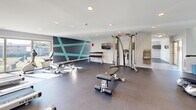 Located on Idlewild Road in Bel Air, Maryland, this single-level fitness center offers a comprehensive workout environment designed to accommodate a wide range of exercise needs. The space is thoughtfully organized into distinct functional zones, allowing users to move seamlessly between cardio training, strength work, and free-weight exercises.

The cardio area is well-appointed with multiple treadmills and elliptical machines, each equipped with individual monitors for an enhanced workout experience. Large windows along the perimeter flood the space with abundant natural light while providing views of the surrounding outdoor greenery, creating an inviting and energizing atmosphere. Wall-mounted televisions are positioned throughout for added convenience.

The strength training section features a cable machine system, a dedicated dumbbell rack with a full range of weights, and multiple adjustable benches to support varied workout routines. A standout design element is the bold geometric accent wall rendered in dark charcoal and teal, adding a modern, dynamic aesthetic to the space.

The fitness center is finished with durable rubber flooring throughout, recessed lighting in the white ceiling, and a built-in water fountain station. The open floor plan and generous spacing between equipment ensure a comfortable and functional experience for all users.