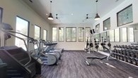This fitness center is designed to provide a comprehensive workout environment within a bright, open-plan layout. The space features elevated ceilings accented by black industrial-style pendant lights and ceiling fans, contributing to a modern, functional aesthetic. The walls are finished in a clean white tone, complemented by large black-framed windows that line multiple walls, flooding the interior with natural light and offering views of surrounding trees and landscaping.

The flooring consists of durable, dark-toned carpet tiles that define the workout area and provide cushioning underfoot. Along one wall, full-length mirrors visually expand the space and allow users to monitor their form during exercise.

The cardio section is well-stocked with multiple treadmills, elliptical machines, and a stair-climbing machine, all positioned to face the windows. The strength training area includes a full rack of dumbbells, a weight bench, a cable machine, and plate-loaded equipment, providing options for a wide range of resistance exercises. A wall-mounted television is also present. The overall ambiance is clean, modern, and energizing, offering a practical and well-lit environment suited to a variety of fitness routines.