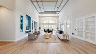Located at 2801 Biltmore Park Dr in Orlando, FL, this single-floor community clubhouse offers a generously proportioned living space designed with a refined, contemporary aesthetic. The centerpiece of the space is a grand open living room distinguished by dramatically soaring ceilings with a decorative coffered ceiling detail and abundant recessed lighting. A striking arched transom window at the far end floods the interior with natural light, creating a bright and welcoming ambiance throughout.

The living area is organized into multiple distinct seating zones, accommodating both casual lounging and social gatherings. Neutral tones of white and warm gray dominate the palette, accented by bold teal-painted built-in display niches along one wall that add a vibrant, curated design element. Light-toned hardwood-style flooring flows continuously across the entire space, enhancing its sense of openness.

White-framed French doors with glass paneling connect seamlessly to adjacent rooms and exterior access points, including a dedicated business center lounge area visible through a full-height glass partition. A dining area with deep navy upholstered chairs and a separate lounge with soft bouclé seating further enrich the variety of functional zones. Lush greenery and thoughtfully placed decor add warmth and life to this polished, community-focused gathering space.