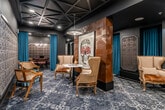 Located in downtown Santa Ana, CA, this single-floor venue is divided into two distinct areas that together create an immersive, lounge-like atmosphere with a strong design identity. The space is defined by deep charcoal walls, a striking geometric coffered ceiling with exposed structural detailing, and a patterned carpet that anchors the eclectic furnishings throughout.

The main lounge area centers around a prominent copper-tiled pillar, which serves as a dramatic architectural focal point. Surrounding it are elegantly mismatched wingback and cane chairs arranged around a marble-topped bistro table, evoking a sophisticated yet intimate gathering environment. A bold skull artwork adorns the pillar, reinforcing the space's distinctive, avant-garde aesthetic. Embossed decorative wall panels add texture throughout, while teal drapery softens transitions between zones. A built-in bar with mirrored tile backsplash and under-counter shelving is tucked along one wall, completing the lounge experience.

The secondary room is dedicated to gaming, featuring a full-sized poker table surrounded by cushioned chairs beneath a candelabra-style chandelier. Octagonal embossed wall panels frame this room with a theatrical, salon-like quality. Together, the two areas offer a cohesive yet layered environment ideal for private social gatherings or entertainment.