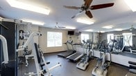 This fitness center is a dedicated, single-floor exercise facility designed to accommodate a well-rounded workout routine. The space is outfitted with multiple treadmills and elliptical machines arranged in a row, providing ample cardio equipment for simultaneous use. A strength training station is positioned along one side, offering additional options for resistance-based exercise. The room features durable, dark rubber flooring suited for high-traffic athletic use, providing both safety and functionality underfoot. Two ceiling fans are mounted overhead, ensuring consistent air circulation throughout the space during workouts. Recessed and flush-mounted fluorescent lighting brightens the room evenly, creating a clear and energetic environment. A wall-mounted flat-screen television is positioned at the front of the room, offering entertainment during exercise sessions. A large mirror spans one wall, visually expanding the space and allowing users to monitor their form. A window along the back wall allows natural light to filter in, adding to the open and airy feel of the room. The neutral gray walls and clean layout contribute to a focused, no-frills atmosphere ideal for fitness.