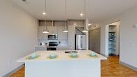 This single-floor unit located on Hobbs Road in Greensboro, North Carolina, presents a clean, contemporary design with a thoughtful open layout. The kitchen serves as a central highlight, showcasing gray shaker-style cabinetry that extends to the ceiling, a white quartz countertop island with an undermount sink, and stainless steel appliances including a refrigerator, range, and microwave. Three pendant lights with glass shades hang above the island, providing both functional lighting and a polished aesthetic. A white subway tile backsplash adds a classic touch to the modern kitchen design.

The living area flows seamlessly from the kitchen, featuring warm-toned hardwood-style flooring and a ceiling fan, creating an open and airy atmosphere. A sliding glass door allows natural light to fill the space and provides access to the outdoors. The bedroom offers plush carpeting, a large window with views of surrounding greenery, and a ceiling fan for added comfort. The bathroom is well-appointed with gray cabinetry, a tub-shower combination with tiled surround, and wood-look flooring. Throughout the unit, neutral tones and consistent finishes create a cohesive, modern living environment.