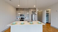 This single-floor unit located on Hobbs Road in Greensboro, North Carolina, presents a clean, contemporary design with a thoughtful open layout. The kitchen serves as a central highlight, showcasing gray shaker-style cabinetry that extends to the ceiling, a white quartz countertop island with an undermount sink, and stainless steel appliances including a refrigerator, range, and microwave. Three pendant lights with glass shades hang above the island, providing both functional lighting and a polished aesthetic. A white subway tile backsplash adds a classic touch to the modern kitchen design.

The living area flows seamlessly from the kitchen, featuring warm-toned hardwood-style flooring and a ceiling fan, creating an open and airy atmosphere. A sliding glass door allows natural light to fill the space and provides access to the outdoors. The bedroom offers plush carpeting, a large window with views of surrounding greenery, and a ceiling fan for added comfort. The bathroom is well-appointed with gray cabinetry, a tub-shower combination with tiled surround, and wood-look flooring. Throughout the unit, neutral tones and consistent finishes create a cohesive, modern living environment.