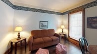 This two-floor space offers a cohesive, comfortable layout distinguished by a recurring decorative floral wallpaper border that ties each room together visually. Wood-trimmed windows fitted with horizontal blinds are found throughout, allowing natural light to filter into every area while maintaining privacy.

The sleeping quarters include multiple bedrooms, each carpeted in deep green and furnished with full or queen beds dressed in blue-and-white striped bedding. Wood nightstands, table lamps, and framed artwork contribute to a cozy, lodge-like atmosphere. One bedroom features a built-in kitchenette nook with a compact refrigerator, microwave, and small sink, along with a wooden desk and office chair — a practical arrangement for extended stays.

The living area is anchored by a plush brown sofa, patterned ottomans, and a flat-screen TV on a wooden media stand, all set atop rich hardwood flooring. A small mirror mounted on the wall adds depth to the room.

The kitchen is functional and well-equipped, featuring wood cabinetry, a gas range, sink, microwave, and countertop appliances. A wood dining table with chairs sits nearby, offering a casual space for meals. The consistent use of warm wood tones, green carpet, and decorative borders throughout gives the entire space a unified, welcoming character.