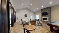 This thoughtfully designed single-floor space makes an immediate impression with its dramatic vaulted ceilings, which lend a sense of openness and grandeur to the main gathering area. Recessed lighting is strategically placed throughout the ceiling, ensuring the space is well-lit and inviting at any time of day. The warm, light-toned hardwood-style flooring runs throughout, tying the design together with a cohesive and clean aesthetic.

The focal point of the room is a floor-to-ceiling stacked stone fireplace, topped with a wall-mounted flat-screen television — a striking combination that anchors the seating area and adds a rustic yet refined character. Neutral-toned walls complement the natural textures found throughout the space.

Furnishings follow a modern farmhouse style, featuring a large sectional sofa with accent pillows, a chunky wood coffee table, and a mix of woven and upholstered accent chairs. A round bistro-style table with black metal chairs near the entry offers a more casual seating option, ideal for conversation or light activity. A decorative sunburst wall accent adds an artistic touch to the neutral backdrop. Natural light filters in through the windows, enhancing the warm and welcoming ambiance that defines this versatile, communal space.