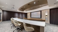 This professionally designed presentation and screening space combines high-end aesthetics with functional versatility. The main area features a tiered layout with curved rows of rich cognac leather armchairs arranged in a theater-style configuration, each row elevated to ensure clear sightlines throughout the room. Warm wood-capped dividers separate the rows, while integrated step lighting adds both safety and ambiance. A ceiling-mounted projector serves the space, supported by a presentation counter at the front with light wood paneling and a built-in ledge beneath the large projection screen. The ceiling is a standout architectural element, featuring a dramatic curved soffit with recessed lighting that adds depth and a sense of refinement to the overall design.

A secondary conference area sits adjacent, furnished with a sleek white elongated table and patterned upholstered task chairs. Floor-to-ceiling dark drapery panels line the walls, providing a polished, controlled environment ideal for focused presentations or screenings. Contemporary abstract artwork adorns the walls of the main seating area, contributing to the space's sophisticated character. Textured carpet flooring in a muted grey tone grounds the design, tying together the warm leather tones and neutral palette throughout. The space is well-suited for corporate presentations, private screenings, or executive briefings.