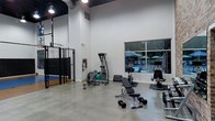 This fitness facility is designed to support a wide range of workout routines within a spacious, single-floor layout. The interior features polished concrete floors and crisp white walls that contribute to a clean, functional aesthetic. Soaring ceilings with exposed industrial-style ductwork and overhead lighting create an open, airy atmosphere throughout the space.

The main workout area is outfitted with a selection of strength training equipment, including adjustable benches, a multi-function cable machine, and a collection of free weights and dumbbells neatly arranged for easy access. Cardio machines are positioned near a large window that overlooks an outdoor area, providing natural light and pleasant views of what appears to be a pool and patio space with umbrella seating.

A separate enclosed training zone is visible behind a black chain-link partition, offering a dedicated area for functional fitness, agility drills, or sport-specific training. This sectioned space adds versatility to the facility's overall offerings.

The layout is thoughtfully organized to maximize usable floor space while maintaining clear pathways between equipment. The combination of natural light, high ceilings, and a well-curated equipment selection makes this facility a comprehensive and inviting environment for fitness enthusiasts of all levels.