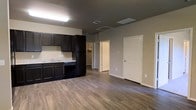 This single-floor space offers a practical and modern open-concept layout that seamlessly integrates the kitchen and living areas. The kitchen is outfitted with dark espresso-toned cabinetry that extends to the ceiling, providing generous storage capacity. Light-colored stone countertops create a striking contrast against the dark cabinets, while a black dishwasher and sink complete the functional workspace. The flooring throughout is a warm, medium-toned hardwood-style plank that adds a sense of continuity and warmth across the entire space. The walls are finished in a neutral gray tone, lending a clean and contemporary aesthetic. Ceiling-mounted fluorescent lighting ensures the space is well-lit, and HVAC vents are thoughtfully integrated into both the ceiling and walls for efficient climate control. A doorway visible from the main area leads to an additional room, suggesting a walk-in closet or utility space, while a classic six-panel white door provides access to another area of the unit. The overall design is minimalist and move-in ready, with clean lines and a neutral palette that offers a versatile backdrop for a variety of interior styles.