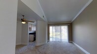 This single-floor residence offers a thoughtfully designed layout that balances comfort and functionality. The living area serves as the heart of the home, featuring plush wall-to-wall carpeting, crown molding, and a ceiling fan that adds both style and practicality. A large sliding glass door dressed with vertical blinds floods the space with natural light while providing access to the outdoors and views of the surrounding greenery, creating a bright and welcoming atmosphere.

Adjacent to the living area, the kitchen is accessible through an open archway, maintaining a sense of connectivity throughout the home. The kitchen is equipped with light wood cabinetry, dark countertops, and includes essential appliances, including a range. The tile flooring in the kitchen offers a clean contrast to the carpeted living space.

Crown molding and clean white trim work run consistently throughout the space, lending a polished, cohesive aesthetic. The neutral color palette of warm beige tones on the walls complements the light-toned finishes, creating an inviting and versatile interior that is easy to personalize. Overall, this residence combines practical design elements with comfortable living spaces, making it a well-rounded and move-in ready home.