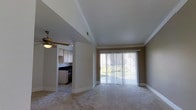 This single-floor residence offers a thoughtfully designed layout that balances comfort and functionality. The living area serves as the heart of the home, featuring plush wall-to-wall carpeting, crown molding, and a ceiling fan that adds both style and practicality. A large sliding glass door dressed with vertical blinds floods the space with natural light while providing access to the outdoors and views of the surrounding greenery, creating a bright and welcoming atmosphere.

Adjacent to the living area, the kitchen is accessible through an open archway, maintaining a sense of connectivity throughout the home. The kitchen is equipped with light wood cabinetry, dark countertops, and includes essential appliances, including a range. The tile flooring in the kitchen offers a clean contrast to the carpeted living space.

Crown molding and clean white trim work run consistently throughout the space, lending a polished, cohesive aesthetic. The neutral color palette of warm beige tones on the walls complements the light-toned finishes, creating an inviting and versatile interior that is easy to personalize. Overall, this residence combines practical design elements with comfortable living spaces, making it a well-rounded and move-in ready home.