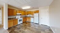 Located at Friendship Village in Harrington, Delaware, this single-floor residence offers two bedrooms and two full bathrooms within a thoughtfully laid-out floor plan. The kitchen is a functional highlight, featuring warm honey-toned oak cabinetry, dark laminate countertops, wood-look vinyl plank flooring, and a full suite of white appliances including a range, dishwasher, and refrigerator. Ample upper and lower cabinet storage makes the space practical for everyday living. Adjacent to the kitchen is a dining area with matching flooring, providing a seamless transition between spaces. The living room is generously proportioned with plush wall-to-wall carpeting and a sliding glass door that invites natural light and opens to an outdoor view. The bedrooms are carpeted and finished with neutral tones; the primary bedroom is notably spacious and includes access to a dedicated closet with wire shelving. Both bathrooms are clean and functional, each featuring a tub-shower combination, a vanity with oak cabinetry, and wood-look tile flooring. A connecting hallway links the private and shared areas of the home. Throughout, neutral beige walls and consistent finishes create a cohesive, move-in-ready atmosphere.