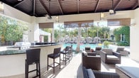 This single-story covered outdoor lounge and bar area offers a seamlessly integrated resort-style experience. The space features a striking wood-paneled ceiling with ceiling fans and pendant lighting, providing shade and comfort while maintaining an open-air atmosphere. A built-in bar with a dark countertop and wicker bar stools anchors one side of the space, ideal for poolside service. The lounge area is furnished with dark wicker seating and cushioned chairs arranged to maximize views of the sparkling pool beyond. Bamboo roll-up shades line the open perimeter, offering adjustable privacy and sun control without obstructing the views. The flooring consists of light-toned tile that extends naturally toward the pool deck. Through the open sides of the structure, a resort-style pool is prominently visible, flanked by rows of lounge chairs and white fabric cabanas, with mature palm trees providing a lush tropical backdrop. The overall ambiance is sophisticated yet relaxed, combining functional amenities with an inviting, vacation-inspired aesthetic well-suited for both casual lounging and social gatherings.
