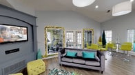 This single-floor community clubhouse offers a well-rounded amenity space designed with a cohesive and energetic color palette of teal, chartreuse green, and cool gray throughout. The main lounge area is generously proportioned, featuring soaring vaulted ceilings, large pendant light fixtures, and abundant natural light streaming through multiple French doors and an arched window. Comfortable seating — including a plush gray velvet sofa accented with teal throw pillows and a tufted chartreuse ottoman — creates an inviting gathering space centered around a wall-mounted flat-screen television. Hardwood-style flooring flows seamlessly throughout the space. An adjacent kitchenette features a curved granite-topped bar with stylish black bar stools, a stainless steel refrigerator, and a microwave, set against a striped accent wall. A dedicated workspace area with a modern desk and office chair provides a functional setting for residents. The fitness center, accessed through French doors adorned with large mirrors, is clearly visible from the lounge and equipped with cardio machines. The space is bright, open, and thoughtfully designed to serve as a versatile hub for socializing, working, and staying active.