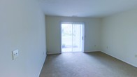 This single-floor space offers a straightforward and functional layout designed for comfortable everyday living. The main living area is finished with wall-to-wall neutral carpeting that lends a warm, understated feel to the interior. A prominent sliding glass door serves as a central architectural feature of the room, fitted with vertical blinds that allow for adjustable light control. This door floods the space with abundant natural light and provides direct access to an outdoor area, offering views of surrounding greenery. The walls are finished in a clean, soft neutral tone that complements the light palette of the room and enhances the sense of openness. The ceiling features a subtle slope, adding a modest architectural detail to the otherwise clean lines of the space. The room's open layout provides flexibility for a variety of furniture arrangements and functional uses. Overall, the space presents a bright, airy ambiance with a practical design, making it a versatile and welcoming environment. The combination of natural light, outdoor access, and neutral finishes creates a calm and livable atmosphere throughout.