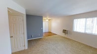This single-floor residence offers a practical and comfortable layout with a notably open living area. The main living space features a bold slate-blue accent wall that adds a touch of visual interest against the neutral white walls and ceiling. Soft, wall-to-wall carpeting covers the living area floor, providing a warm underfoot feel throughout the room.

A ceiling fan is centrally positioned to ensure comfortable air circulation year-round, complementing the wall-mounted HVAC unit installed along one wall, offering convenient climate control. Natural light filters in through a side window, brightening the interior and enhancing the sense of openness.

The living space transitions into an adjoining area with hardwood-style flooring, partially visible beyond the accent wall partition, adding a subtle contrast in materials and defining distinct zones within the home. Vertical blinds at the entryway offer privacy and light control at the threshold.

White interior doors and clean baseboards frame the space with a crisp, tidy finish. The overall ambiance is functional and uncluttered, making it an easily adaptable canvas for a variety of furnishing styles. The straightforward, single-floor layout ensures seamless flow and accessibility throughout the entire space.
