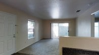 This single-story space features an open-concept layout that connects the living area to the kitchen. The living room is finished with wall-to-wall carpet and neutral beige walls, creating a clean, understated backdrop. A textured popcorn ceiling adds a classic touch overhead, while a side window and a full-width sliding glass door flood the room with natural light and provide direct access to an enclosed outdoor patio area. The sliding door offers views of a fenced exterior space, enhancing the sense of connection between indoors and outdoors. A partial kitchen counter with a dark granite or laminate surface is visible at the edge of the living area, indicating a seamless transition between the two spaces. Ceiling-mounted HVAC vents suggest a central air system throughout. The overall ambiance is bright, open, and functional, suited for everyday living with a practical, no-frills design aesthetic.