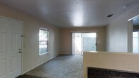 This single-story space features an open-concept layout that connects the living area to the kitchen. The living room is finished with wall-to-wall carpet and neutral beige walls, creating a clean, understated backdrop. A textured popcorn ceiling adds a classic touch overhead, while a side window and a full-width sliding glass door flood the room with natural light and provide direct access to an enclosed outdoor patio area. The sliding door offers views of a fenced exterior space, enhancing the sense of connection between indoors and outdoors. A partial kitchen counter with a dark granite or laminate surface is visible at the edge of the living area, indicating a seamless transition between the two spaces. Ceiling-mounted HVAC vents suggest a central air system throughout. The overall ambiance is bright, open, and functional, suited for everyday living with a practical, no-frills design aesthetic.