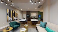 Located at 1302 Rio Valley Dr in Raleigh, NC, this single-story community clubhouse offers a thoughtfully designed gathering space that balances modern aesthetics with welcoming comfort. The entry lobby sets an immediate tone with soaring double-height ceilings, floor-to-ceiling black-framed glass doors and windows that flood the space with natural light, and a distinctive reception desk finished in a rich green textured surface, accented by a striking cluster of geometric gold pendant lights overhead. Wide-plank wood-look flooring flows consistently throughout, creating visual continuity across all areas. The open lounge transitions seamlessly into a communal dining and social zone, where marble-topped tables with gold-accented bases are paired with upholstered dining chairs, complemented by bold abstract wall art in gold, black, and navy tones. Emerald green velvet accent chairs add a vibrant focal point to the seating area. A kitchenette and bar area with dark cabinetry and subway tile backsplash serves the social spaces. A curved media lounge anchors one end of the clubhouse, wrapped in deep green textured wall covering, built-in window bench seating, brass cylindrical side tables, and a tufted ottoman beneath a sculptural brass multi-globe chandelier, with large windows framing outdoor greenery views.