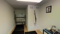 This single-floor pet grooming facility is thoughtfully designed to support professional grooming operations in a clean, functional environment. The space features light-toned walls that create a bright and neutral backdrop, complemented by hardwood-style flooring throughout. Anti-fatigue rubber mats are strategically placed in key work areas to provide comfort during extended grooming sessions and to maintain safety on potentially wet surfaces. At the heart of the facility is a professional-grade stainless steel grooming tub, positioned within a dedicated grooming corridor that keeps the workflow organized and efficient. The tub is equipped with the necessary fixtures to handle bathing and grooming tasks with ease. A grooming apron hangs conveniently on the wall, keeping essential gear accessible and ready for use. Ventilation is well-addressed with multiple HVAC vents installed both on the ceiling and along the lower walls, ensuring proper air circulation throughout the space. A suspended ceiling with integrated lighting provides consistent, even illumination across the work area. The overall layout is compact yet purposefully arranged, making it a practical and well-equipped environment suited for pet grooming professionals.