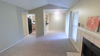 This single-floor residence showcases a thoughtfully designed open-concept layout that creates a seamless flow between the main living areas. The living room is a standout feature, boasting dramatic vaulted ceilings that lend a sense of volume and openness to the space. Soft, neutral-toned carpeting covers the floors, contributing to a warm and inviting atmosphere throughout. Large windows fitted with blinds allow for generous natural light while maintaining privacy, brightening the interior throughout the day. A pass-through opening connects the living area to the kitchen, which is equipped with wooden cabinetry, a dark countertop, and full-size appliances including a black refrigerator. The kitchen layout provides practical functionality and easy accessibility from the main living space. The neutral color palette used throughout — from the beige walls to the light trim — creates a cohesive and clean aesthetic that is easy to personalize. The open sightlines between rooms enhance the sense of space, making the interior feel expansive despite being contained to a single floor. Overall, this space offers a comfortable and functional living environment with architectural details that elevate its everyday appeal.