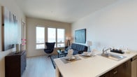 This stylish single-floor residence offers a thoughtfully designed open-concept layout that connects the kitchen, dining, and living areas into one cohesive living environment. The kitchen features a clean white countertop that extends into a dining surface, providing a functional and visually streamlined transition between spaces. The dining area is set for entertaining, with place settings neatly arranged along the countertop bar.

The living area is anchored by a charcoal blue sofa paired with a matching Egg-style accent chair, both set atop a geometric patterned area rug over light hardwood-style flooring. A framed map artwork in deep blue tones adds a sophisticated focal point to the wall, complementing the cool color palette found throughout the space.

Large windows along the far wall flood the interior with abundant natural light and offer expansive views of the surrounding skyline and waterfront, enhancing the airy, open ambiance of the room. The clean white walls and smooth ceilings contribute to a sense of spaciousness and simplicity. Overall, the space presents a modern, well-appointed aesthetic that balances comfort with contemporary design sensibility.