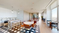 Located at 421 Summit Ave in Jersey City, NJ, this expansive two-floor amenities suite offers a refined, modern retreat designed for both socializing and relaxation. The main floor hosts a combined dining and living area anchored by a striking marble-topped island with bar seating, stainless steel appliances, and bold checkered tile flooring that transitions into warm hardwood. The living area features plush seating arrangements, built-in cabinetry with mirrored accents, and floor-to-ceiling windows that flood the space with natural light and deliver breathtaking panoramic views of the Jersey City skyline and beyond. Intimate café-style seating areas are positioned directly against the expansive glazing to maximize the elevated vistas. A glass-railed staircase with brass pendant lighting connects the two floors with architectural elegance. The upper level houses a well-appointed game room complete with a pool table, shuffleboard table, and casual lounge seating, all bathed in natural light from oversized windows framing wide open city views. The main living area on this floor offers curved, contemporary sofas arranged around a central coffee table, with corner glass walls providing an immersive, panoramic backdrop throughout.