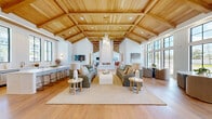 This single-floor space offers a generously sized, open-concept layout defined by striking architectural details and a cohesive coastal-modern aesthetic. The centerpiece of the main living area is a dramatic vaulted ceiling clad in warm natural wood with exposed beam framing, lending the space a grand yet inviting atmosphere. A sculptural floor-to-ceiling white fireplace anchors the central gathering area, complementing the expansive seating arrangements set atop woven rugs over wide-plank oak hardwood floors.

Floor-to-ceiling black-framed windows line multiple walls throughout, flooding the interior with natural light and providing outdoor views. The open kitchen features sleek white cabinetry with brass hardware, a marble-veined stone countertop, stainless steel appliances including a dishwasher and beverage refrigerator, and an oversized undermount sink beneath a row of large windows. Distinctive arched wood-paneled alcoves add warmth and architectural character near the kitchen.

A dedicated office area features textured statement wallcovering, globe wall sconces, and a built-in floating desk. An additional flex room provides versatile functionality. Together, the rooms create a bright, airy, and well-appointed space with refined coastal charm throughout.