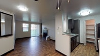 This single-floor residence offers a thoughtfully designed open-concept layout that seamlessly connects the kitchen and living areas. The living room is anchored by a classic tiled fireplace with a white mantel, creating a welcoming focal point. Rich, dark hardwood-style flooring runs throughout, lending warmth and continuity to the space. A sliding glass door with vertical blinds fills the living area with natural light while providing direct access to the outdoors. The kitchen features dark-stained cabinetry with brushed hardware, a countertop peninsula with an integrated sink, and pendant lighting overhead — a practical yet stylish design. A half-wall partition elegantly separates the kitchen from the living space, preserving an open feel while defining distinct functional zones. The neutral color palette of soft grays and whites on the walls complements the darker tones of the cabinetry and flooring, resulting in a balanced and modern aesthetic. Ceiling-mounted flush lighting and a visible thermostat panel reflect the space's attention to everyday convenience. Overall, this residence combines functional design with contemporary finishes, making it a comfortable and inviting single-level living environment.